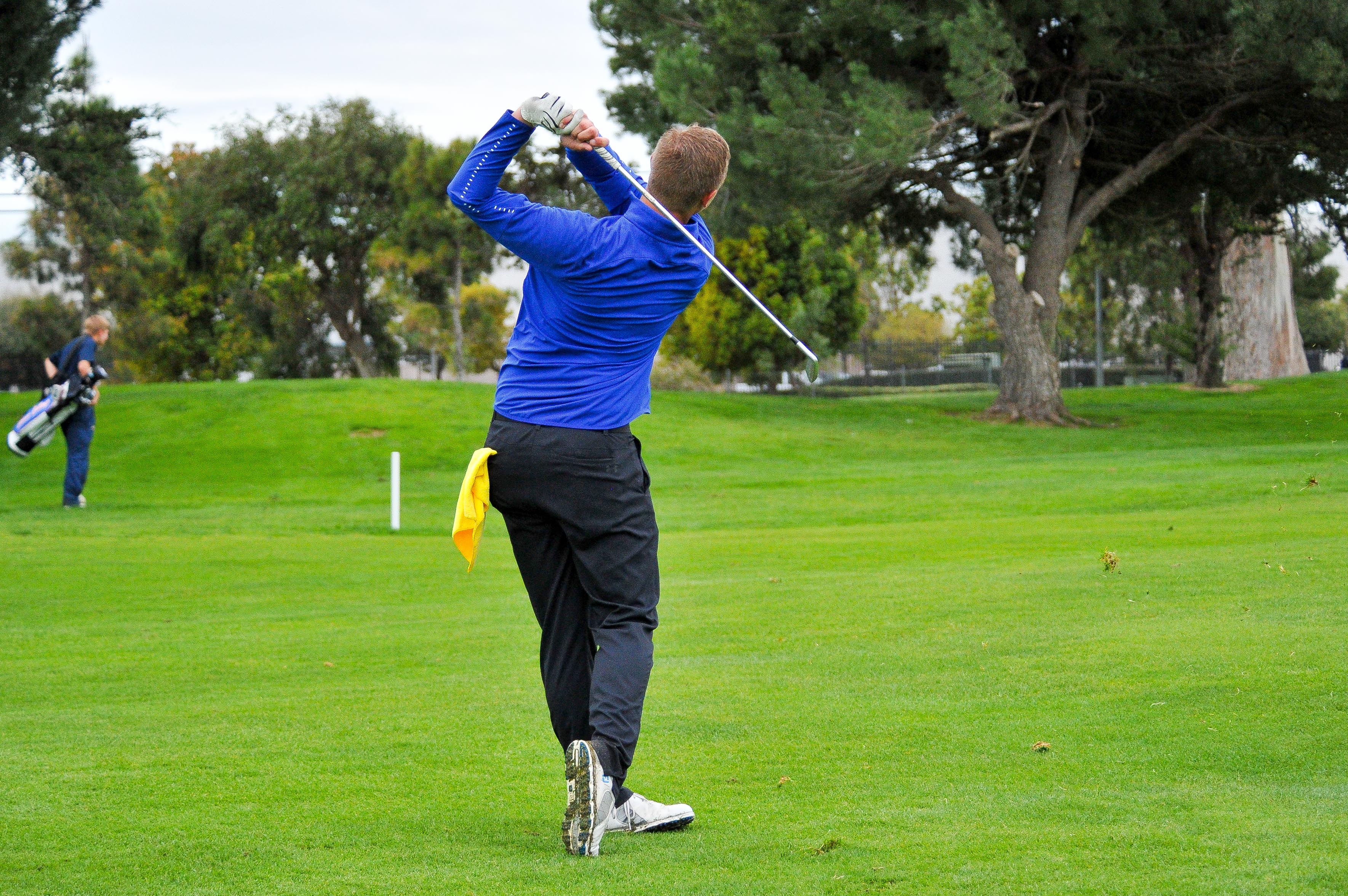 A golfer swings a club on a green fairway during Allan Hancock College Athletics’ Final Fore Golf Tournament. The golfer, wearing a blue long-sleeve shirt and black pants, follows through on the shot while another participant with a golf bag walks in the background on the course.